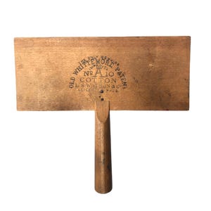 May include: A vintage wooden cotton smoother with a rectangular head and a handle. The head features the text "OLD WHITTEMORE PATENT" and "NO. A 10 COTTON". The tool is made of light brown wood.