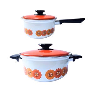 May include: Two vintage cooking pots with lids. The pots are white with orange floral patterns and orange lids. One pot has a black handle, and the other has two black side handles. The lids have black knobs.