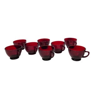 May include: A set of eight ruby red glass teacups. Each cup has a rounded bowl shape, a small handle, and a dark base. The cups are arranged in a slightly curved line against a white background, showcasing their rich color and design.