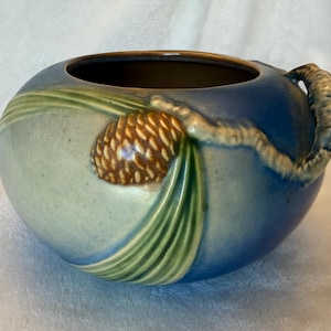 May include: A round ceramic bowl featuring a gradient of blue and light green glaze. The exterior is decorated with a raised, molded relief of a brown pinecone attached to a textured branch and long, slender green pine needles.