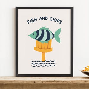 Könnte beinhalten: Ein gerahmter Druck mit der Aufschrift "FISH AND CHIPS" über einem Cartoon-Fisch mit blau-weißen Streifen. Der Fisch sitzt auf einer gelben Struktur über blauen Wellen. Ein Salzstreuer und ein Teller Pommes liegen auf einer Holzoberfläche.