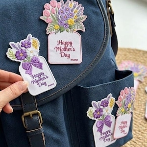 Könnte beinhalten: Eine marineblaue Canvas-Tasche mit mehreren gestickten Aufnähern. Die Aufnäher zeigen Blumensträuße in Vasen mit dem Text "Happy Mother's Day". Die Aufnäher sind in Lila, Rosa und Gelb gehalten. Der Text "IRON ON PATCH CUSTOM ANY REQUESTS" ist unten zu sehen.