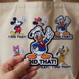 Könnte beinhalten: Beige Tragetasche mit Aufnähern von Zeichentrickfiguren wie Mickey Mouse, Minnie Mouse, Donald Duck, Daisy Duck und Pluto. Jeder Aufnäher zeigt den Schriftzug "I DID THAT!" oder "DID THAT!" in schwarzer Schrift.