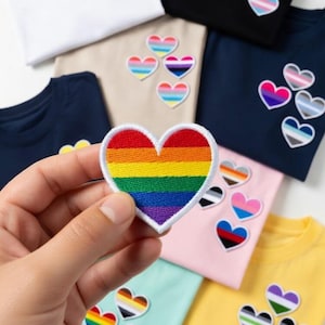 May include: A hand holding a rainbow heart-shaped patch with a white border. Several t-shirts in various colors, including white, black, navy, and yellow, are visible, each adorned with heart-shaped patches in different pride flag designs.
