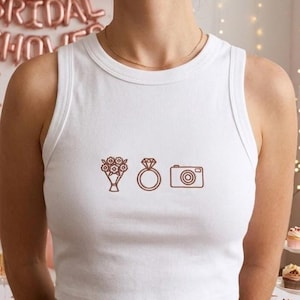 May include: A white sleeveless crop top featuring three embroidered brown icons centered on the chest: a bouquet of flowers, a diamond engagement ring, and a camera. The garment has a ribbed crew neckline and a fitted silhouette.