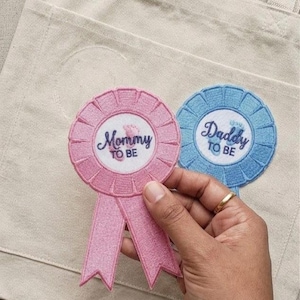 May include: Pink and blue embroidered rosettes with the text "Mommy to Be" and "Daddy to Be". The pink rosette has a pink ribbon. The rosettes are held in front of a beige tote bag.