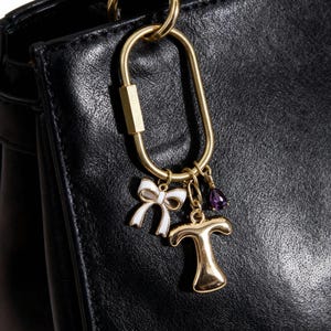 Könnte beinhalten: Nahaufnahme einer schwarzen Lederhandtasche mit einem goldfarbenen Karabinerhaken. Am Haken hängen ein weißer Schleifenanhänger, ein goldener Buchstabe "T" und ein lila Edelsteinanhänger. Die Tasche hat einen goldenen Griff.