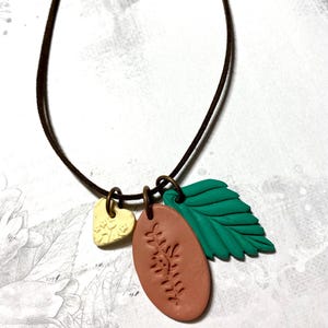 May include: A necklace with a brown cord and three charms. A small, cream-coloured heart, a reddish-brown oval with a leaf design, and a green leaf. The charms are connected with bronze-coloured rings.