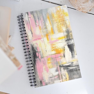 May include: Spiral-bound notebook with an abstract art cover. The cover features a mix of pink, yellow, gray, and black brushstrokes, creating a textured effect. The notebook's spiral binding is black.