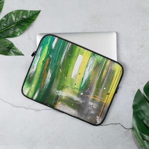 Könnte beinhalten: Laptoptasche mit einem abstrakten Design in Grün-, Gelb-, Weiß- und Grautönen. Die Kunst zeigt vertikale Pinselstriche und einen schwarzen Reißverschluss. Die Hülle liegt auf einem silbernen Laptop.