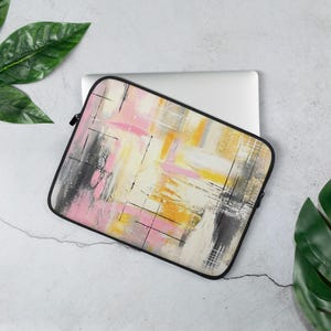 Peut inclure: Housse d'ordinateur portable avec un motif abstrait rose, jaune, noir et blanc. La housse a une fermeture éclair noire et un bord noir. Le motif présente des coups de pinceau et des lignes entrecroisées. La housse est sur un ordinateur portable blanc.