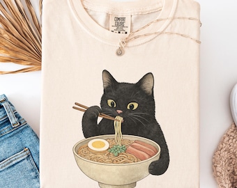May include: Ivory-colored t-shirt featuring a black cat eating ramen from a bowl. The cat is holding chopsticks and the bowl contains noodles, egg, and meat. The shirt has the text "COMFORT COLORS" on the tag. The color of the shirt is ivory.