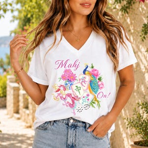 May include: A white V-neck t-shirt featuring a colorful graphic print of a peacock, a pink dragon, flowers, and mahjong tiles. The text "Mahj On!" is printed in pink script above and to the side of the central illustration on the front of the shirt.