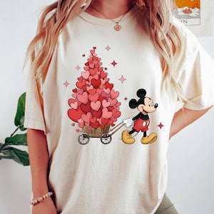Puede incluir: Camiseta color marfil con un dise&ntilde;o de Mickey Mouse tirando de un carro lleno de decoraciones en forma de coraz&oacute;n. El dise&ntilde;o incluye un &aacute;rbol hecho de corazones y acentos de estrellas rojas, ideal para San Valent&iacute;n. El texto "COLOR: IVORY" est&aacute; en la parte inferior.