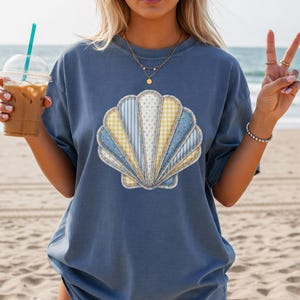 May include: A blue t-shirt featuring a seashell design. The seashell is made of various fabrics in shades of blue and yellow. The person is holding a beverage with a straw.