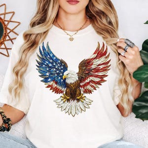 May include: Cream-colored t-shirt featuring a detailed graphic of an American bald eagle with wings spread. The eagle's wings are designed with the American flag's colors and stars. The eagle's body is brown, with a white head and yellow beak.