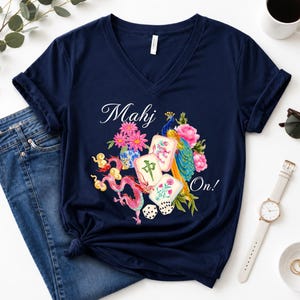 May include: A navy blue V-neck t-shirt featuring a graphic print of mahjong tiles, a pink dragon, a peacock, and flowers. The text "Mahj On!" is printed in white script. The design includes colorful floral elements and two white dice.