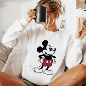 May include: White sweatshirt featuring a classic Mickey Mouse graphic. The cartoon character is depicted in black, red, and white, with a smiling expression. The sweatshirt is a casual, comfortable style.