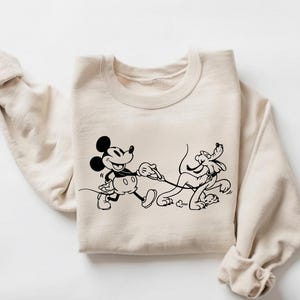 May include: A beige sweatshirt featuring a black graphic of Mickey Mouse walking Pluto on a leash. Mickey is smiling, and Pluto appears to be running. The sweatshirt has long sleeves and a crew neck.