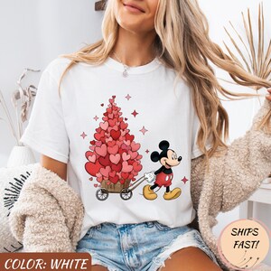 Puede incluir: Camiseta blanca con Mickey Mouse tirando de un carro lleno de decoraciones en forma de coraz&oacute;n. El dise&ntilde;o incluye corazones rosas, estrellas y el texto "COLOR: WHITE". La camiseta tiene cuello redondo y mangas cortas.