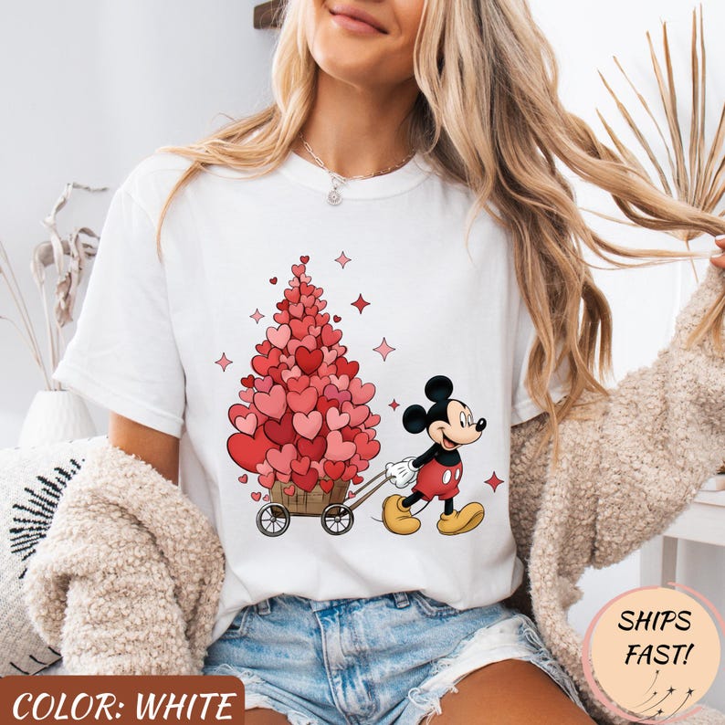 Puede incluir: Camiseta blanca con Mickey Mouse tirando de un carro lleno de decoraciones en forma de coraz&oacute;n. El dise&ntilde;o incluye corazones rosas, estrellas y el texto "COLOR: WHITE". La camiseta tiene cuello redondo y mangas cortas.