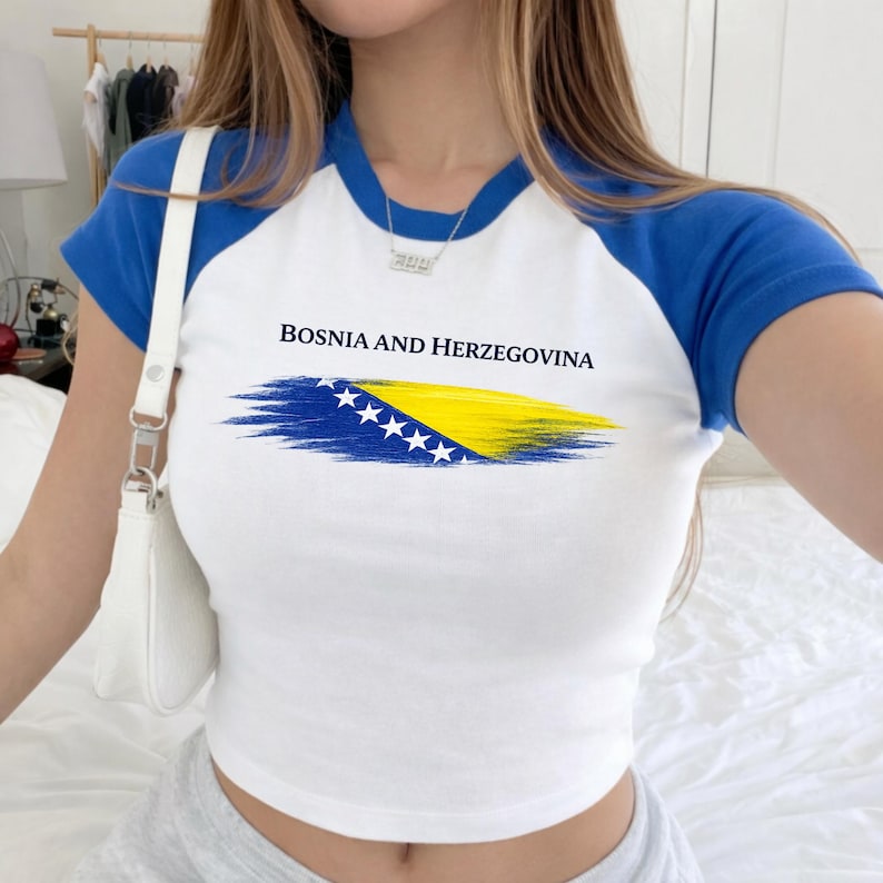 May include: White crop top with blue sleeves and collar, featuring the text "BOSNIA AND HERZEGOVINA" above a painted flag design in blue, yellow, and white. The shirt is a casual style.