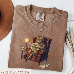 May include: Espresso-colored t-shirt featuring a cat in a suit reading "HOW TO REMOVE A CRIMINAL PRESIDENT." The cat is seated in a red armchair with a lamp and coffee mug on a side table. A silver necklace and a brown hat are also visible.