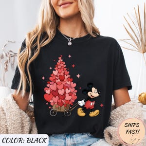 Puede incluir: Camiseta negra con un dise&ntilde;o de Mickey Mouse tirando de un carro lleno de decoraciones en forma de coraz&oacute;n. El texto "COLOR: BLACK" est&aacute; en la parte inferior izquierda y "SHIPS FAST!" en la parte inferior derecha.