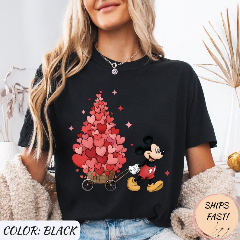 Puede incluir: Camiseta negra con un dise&ntilde;o de Mickey Mouse tirando de un carro lleno de decoraciones en forma de coraz&oacute;n. El texto "COLOR: BLACK" est&aacute; en la parte inferior izquierda y "SHIPS FAST!" en la parte inferior derecha.