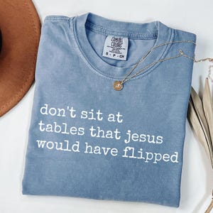 Peut inclure: Un t-shirt bleu Comfort Colors avec le texte blanc « don't sit at tables that jesus would have flipped ». Un collier en or avec un pendentif est drapé sur le t-shirt.