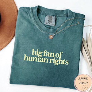 Peut inclure: T-shirt Comfort Colors vert d'eau avec l'inscription jaune « big fan of human rights ». Un collier doré avec un pendentif est posé sur le t-shirt. Le t-shirt est plié sur une surface blanche.