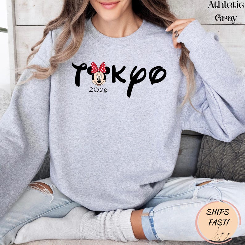 May include: Athletic gray sweatshirt with the word "Tokyo" in black script, featuring a Minnie Mouse graphic with a red bow. The year "2026" is printed below the graphic. The sweatshirt is a light gray color.