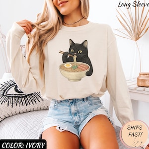 May include: Ivory long-sleeve shirt featuring a black cat eating ramen from a bowl. The cat is holding chopsticks. The shirt has the text "Long Sleeve" at the top and "Color: Ivory" at the bottom.