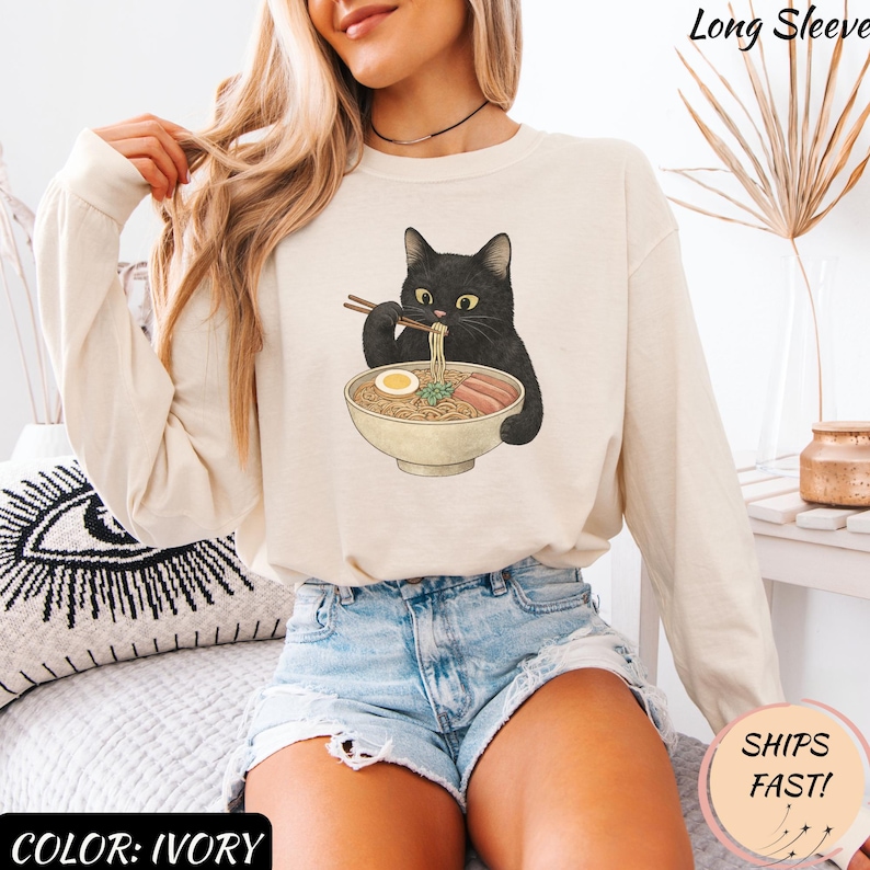 May include: Ivory long-sleeve shirt featuring a black cat eating ramen from a bowl. The cat is holding chopsticks. The shirt has the text "Long Sleeve" at the top and "Color: Ivory" at the bottom.