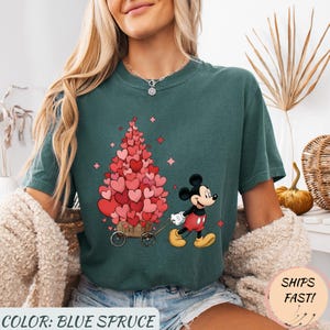 Puede incluir: Camiseta azul abeto con un gr&aacute;fico de Mickey Mouse tirando de un carro lleno de decoraciones en forma de coraz&oacute;n. El dise&ntilde;o incluye corazones rosas y rojos, y estrellas rosas. El texto "COLOR: BLUE SPRUCE" est&aacute; en la parte inferior.