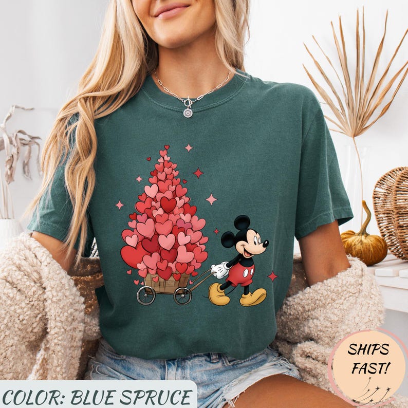 Puede incluir: Camiseta azul abeto con un gr&aacute;fico de Mickey Mouse tirando de un carro lleno de decoraciones en forma de coraz&oacute;n. El dise&ntilde;o incluye corazones rosas y rojos, y estrellas rosas. El texto "COLOR: BLUE SPRUCE" est&aacute; en la parte inferior.