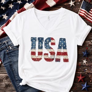Op de afbeelding: Witte V-hals T-shirt met het woord "USA" in een distressed Amerikaans vlagontwerp. De letters zijn blauw met witte sterren en rode strepen. Het shirt ligt op een houten oppervlak met patriottische decoraties.
