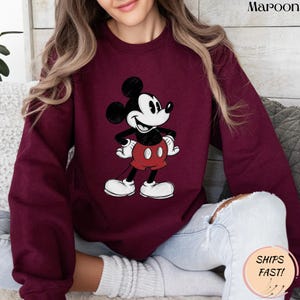 May include: Maroon sweatshirt featuring a classic Mickey Mouse graphic. The cartoon character is depicted in black, white, and red. The sweatshirt has a crew neck and long sleeves. The text "SHIPS FAST!" is on a sticker.