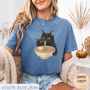 May include: Blue jean colored t-shirt featuring a black cat eating ramen noodles from a bowl. The cat is holding chopsticks and the ramen includes noodles, egg, and meat. The text "COLOR: BLUE JEAN" is at the bottom of the shirt.