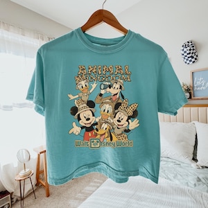 May include: A turquoise t-shirt featuring a graphic of Disney characters, including Mickey Mouse, Donald Duck, and Goofy, in safari attire. The text "ANIMAL KINGDOM" and "Walt Disney World" are printed on the shirt.