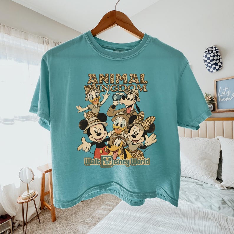 May include: A turquoise t-shirt featuring a graphic of Disney characters, including Mickey Mouse, Donald Duck, and Goofy, in safari attire. The text "ANIMAL KINGDOM" and "Walt Disney World" are printed on the shirt.