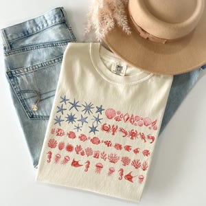May include: A cream-colored t-shirt featuring a graphic print of blue starfish and red seashells, crabs, fish, coral, jellyfish, and stingrays arranged in a flag-like pattern. The shirt is paired with light blue denim jeans, a tan hat, and a necklace.