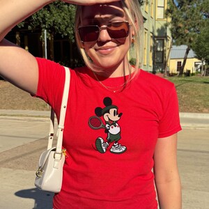 May include: A red t-shirt featuring a cartoon Mickey Mouse playing tennis. Mickey is wearing green shorts, a white shirt, and tennis shoes. The shirt has a small white purse with a gold clasp.