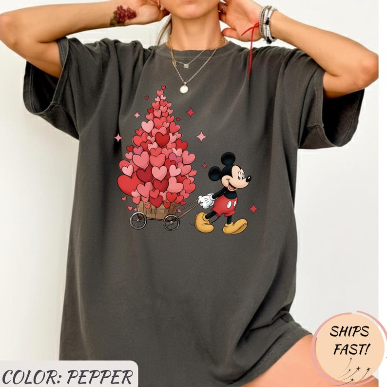 Puede incluir: Camiseta gris oscuro con Mickey Mouse tirando de un carro lleno de decoraciones en forma de coraz&oacute;n. El dise&ntilde;o tiene un tema de San Valent&iacute;n con corazones rosas y rojos. El texto "COLOR: PEPPER" es visible.