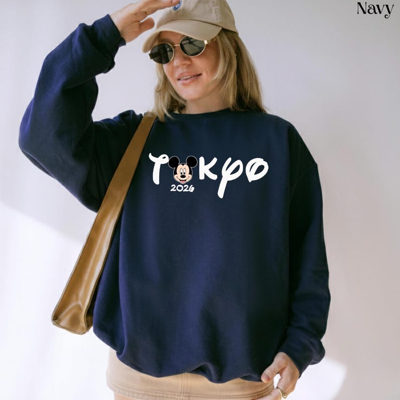 May include: Navy blue sweatshirt featuring the word "Tokyo" in white script, with a Mickey Mouse graphic and the year "2024" below. The sweatshirt is a solid navy color and has a crew neck.