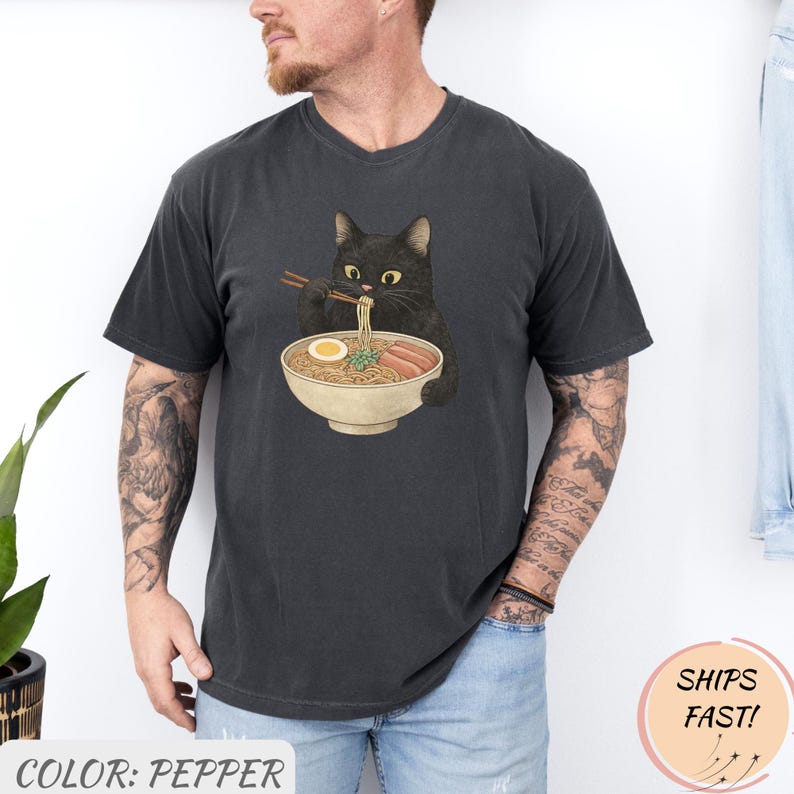 May include: Pepper-colored t-shirt featuring a black cat enjoying ramen. The cat is depicted with chopsticks, eating noodles from a bowl. The shirt has a crew neck and short sleeves. The text "COLOR: PEPPER" is at the bottom.
