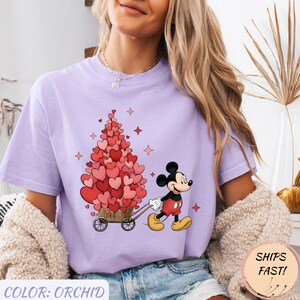 Puede incluir: Camiseta color orqu&iacute;dea con Mickey Mouse tirando de un carro lleno de decoraciones en forma de coraz&oacute;n. El dise&ntilde;o incluye corazones rojos y rosas, con el texto "COLOR: ORCHID" en la parte inferior. La camiseta tiene un corte holgado.