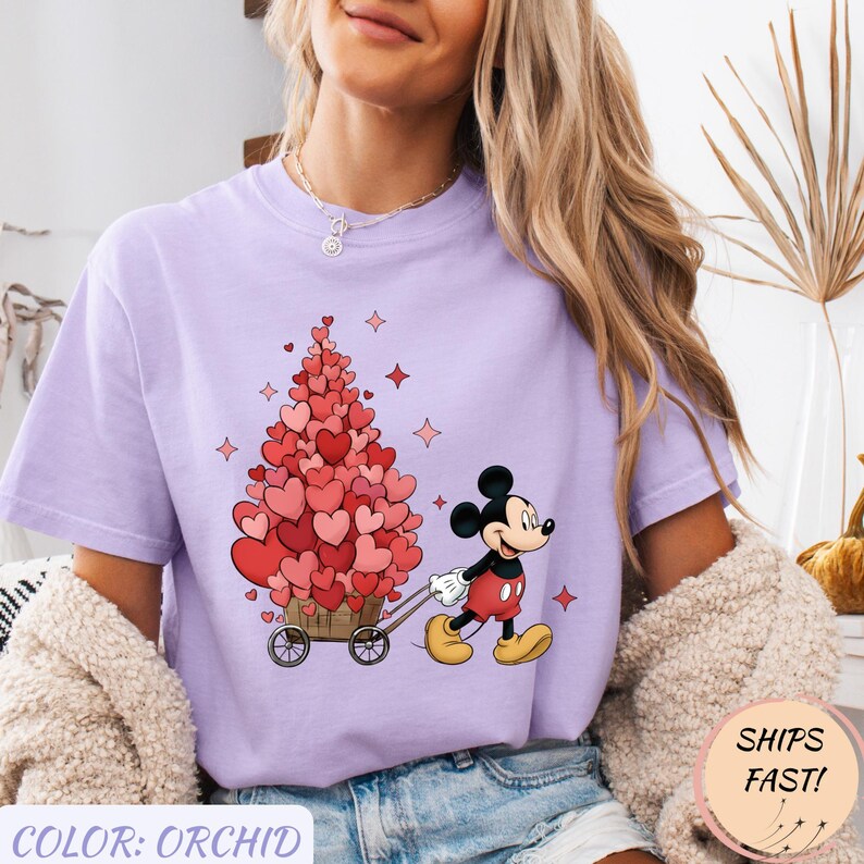 Puede incluir: Camiseta color orqu&iacute;dea con Mickey Mouse tirando de un carro lleno de decoraciones en forma de coraz&oacute;n. El dise&ntilde;o incluye corazones rojos y rosas, con el texto "COLOR: ORCHID" en la parte inferior. La camiseta tiene un corte holgado.