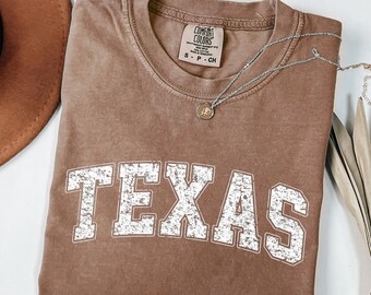 Puede incluir: Camiseta color espresso con la palabra "TEXAS" en letras blancas desgastadas. La camiseta tiene cuello redondo y mangas cortas. Un collar plateado con un colgante está drapeado sobre la camiseta. La camiseta está doblada sobre una superficie blanca.