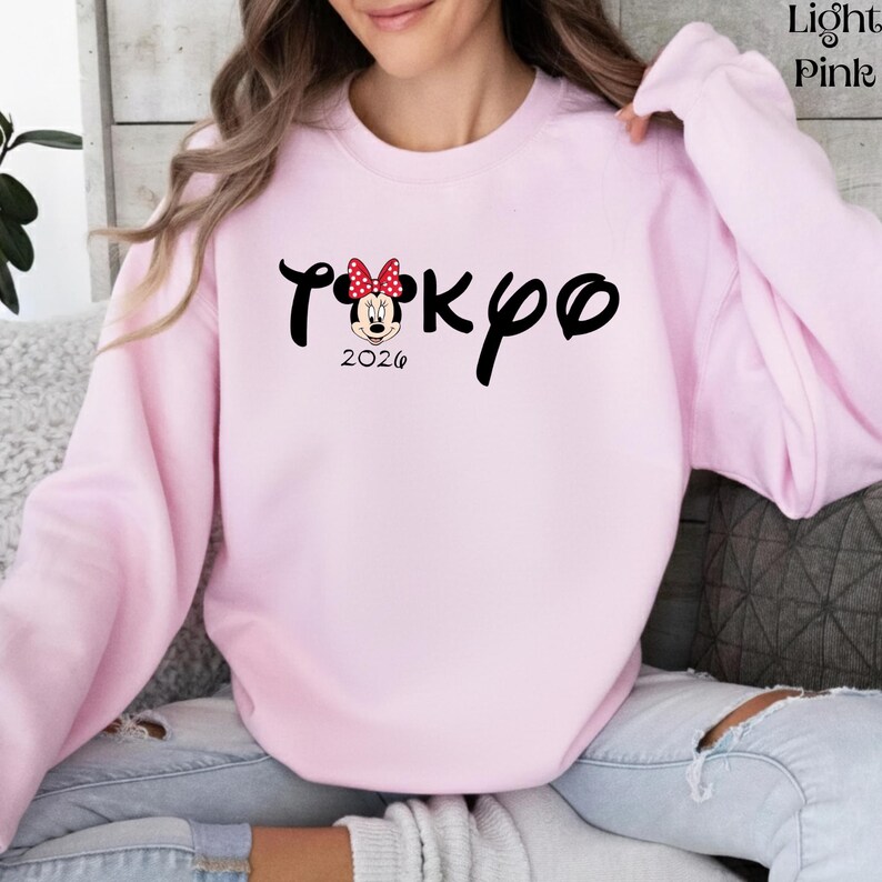 May include: Light pink sweatshirt with the word "Tokyo" in black script, featuring a Minnie Mouse graphic with a red bow. The year "2026" is printed below the graphic. The sweatshirt is a solid light pink color.